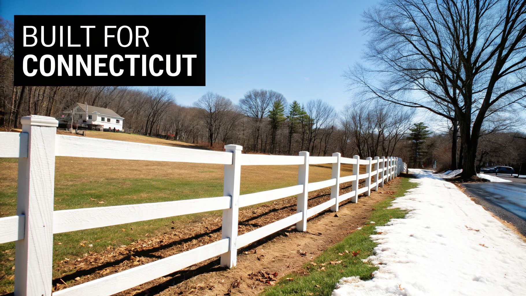 A white post-and-rail fence alongside a snowy road leading to a house on a hill under a blue sky.